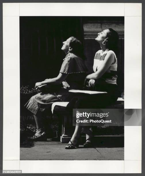 View of two unidentified women seated on a bench, both with their eyes closed and faces tilted toward the sunshine, New York, New York, August 1954.