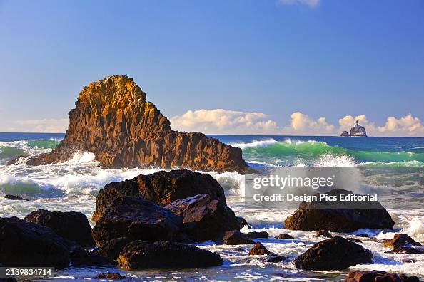 Tillamook Rock Light, a deactivated lighthouse off the coast of Oregon, seen in the distance and waves of the surf rolling into shore and splashing into rocks. Oregon, United States of America