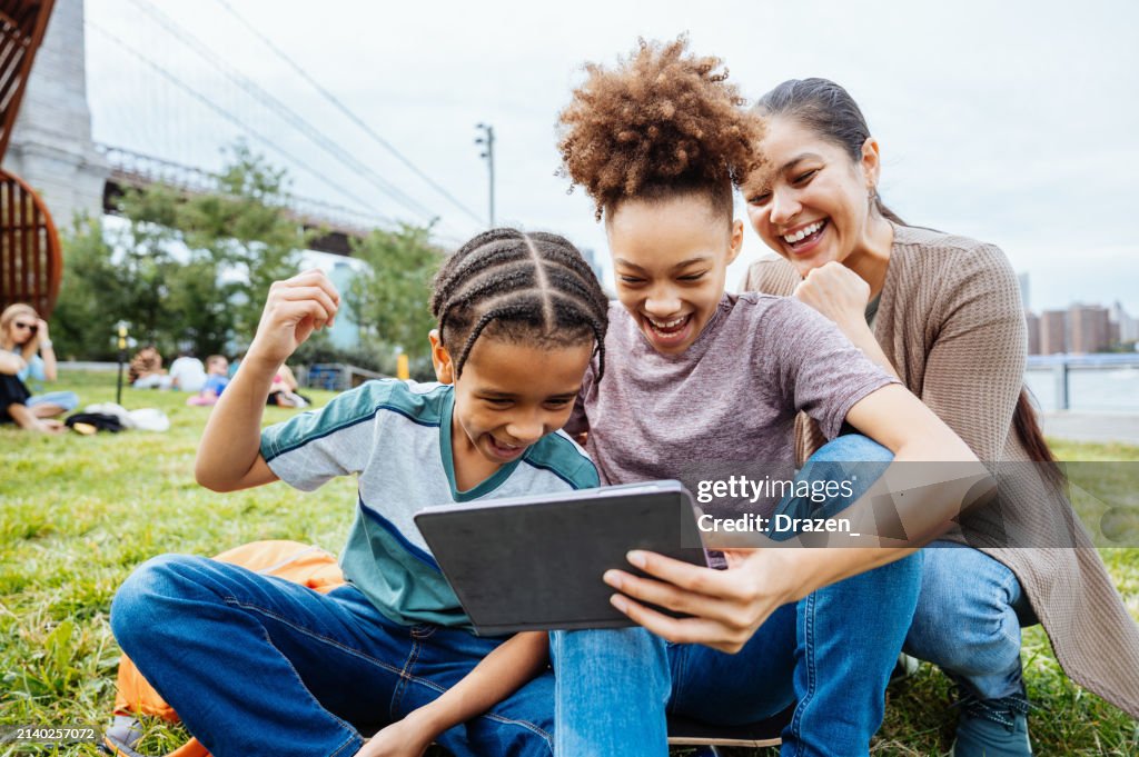 Mid adult mother with two children outdoors. Young teenage siblings using digital tablet to play video games