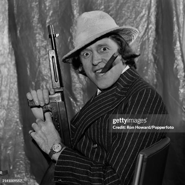 English DJ and television presenter Jimmy Savile posed with large cigar and holding a replica submachine gun backstage in London, circa 1966.