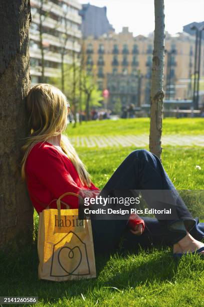 Young Woman Sits Against A Tree on The Grass With A Shopping Bag. Barcelona, Spain.