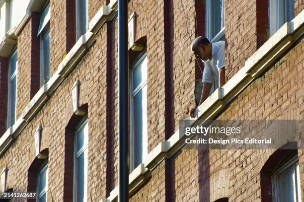 Man Leans Out The Window of A Brick Residential Building Looking Below. London, England.