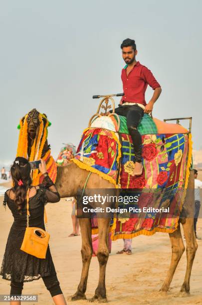 Young Woman taking picture of young man sitting on camel at Puri Beach. Puri, Odisha State, India.