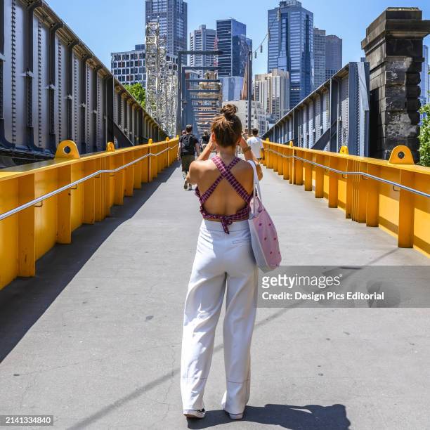 View taken from behind of a woman tourist taking a picture of the Melbourne skyline from a pedestrian bridge. Melbourne, Victoria, Australia.