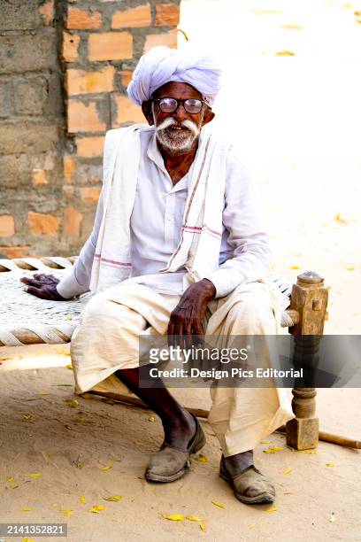 Old man with white turban and moustache sitting on charpoy bed in Bishoi Village. Pali District, Rajasthan, India.