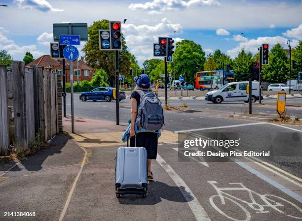Woman pulling suitcase down a sidewalk with a view of traffic in a traffic circle. Oxford, England.