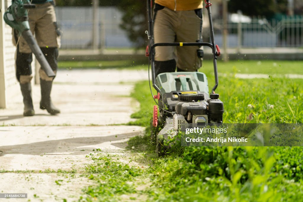 Two men are trimming grass: one is using a lawn mower, and the other one is using a blower to blow away the cut grass.