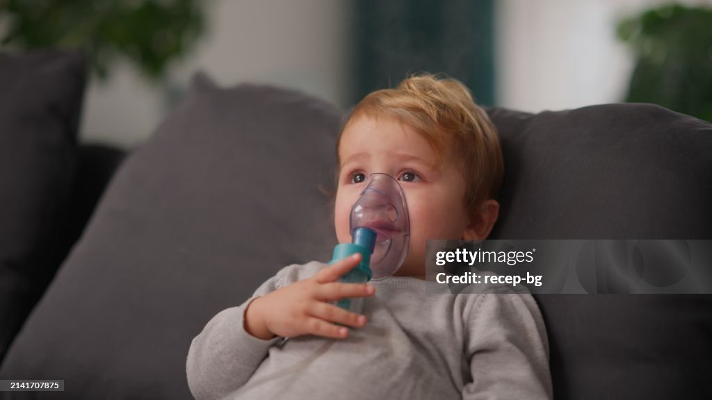 Preschool age boy using nebulizer while sitting on sofa in living room at home