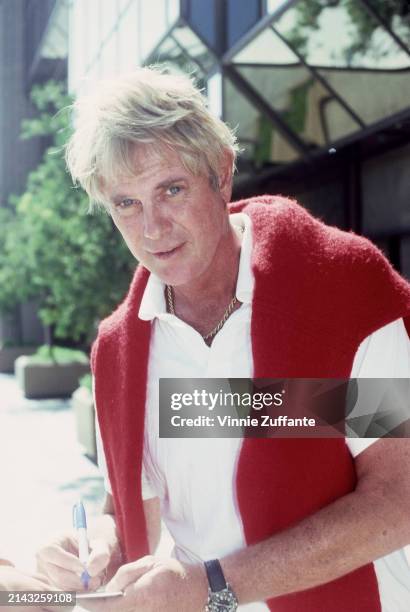 American actor and singer Troy Donahue, wearing a red sweater draped over a white polo shirt, signs an autograph, July 1982.