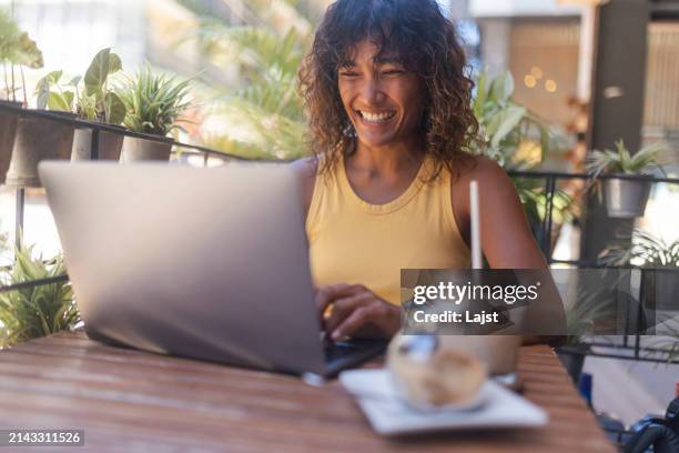 smiling asian woman uses a laptop for remote work while sitting in a coffee shop drinking coffee - digital nomad stock pictures, royalty-free photos & images