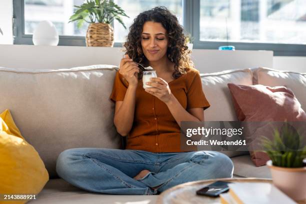 smiling woman eating yogurt on sofa at home - yogurt stock pictures, royalty-free photos & images