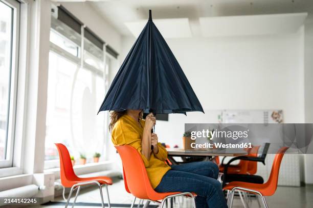 creative businesswoman holding folded umbrella covering face at workplace - verlegen stockfoto's en -beelden