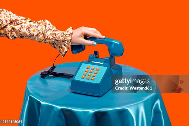 woman picking up telephone receiver on table against orange background - teléfono con cable fotografías e imágenes de stock
