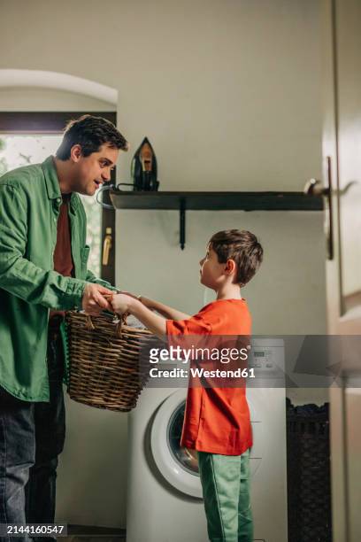 son helping father in holding basket near washing machine at home - man washing basket child stock pictures, royalty-free photos & images