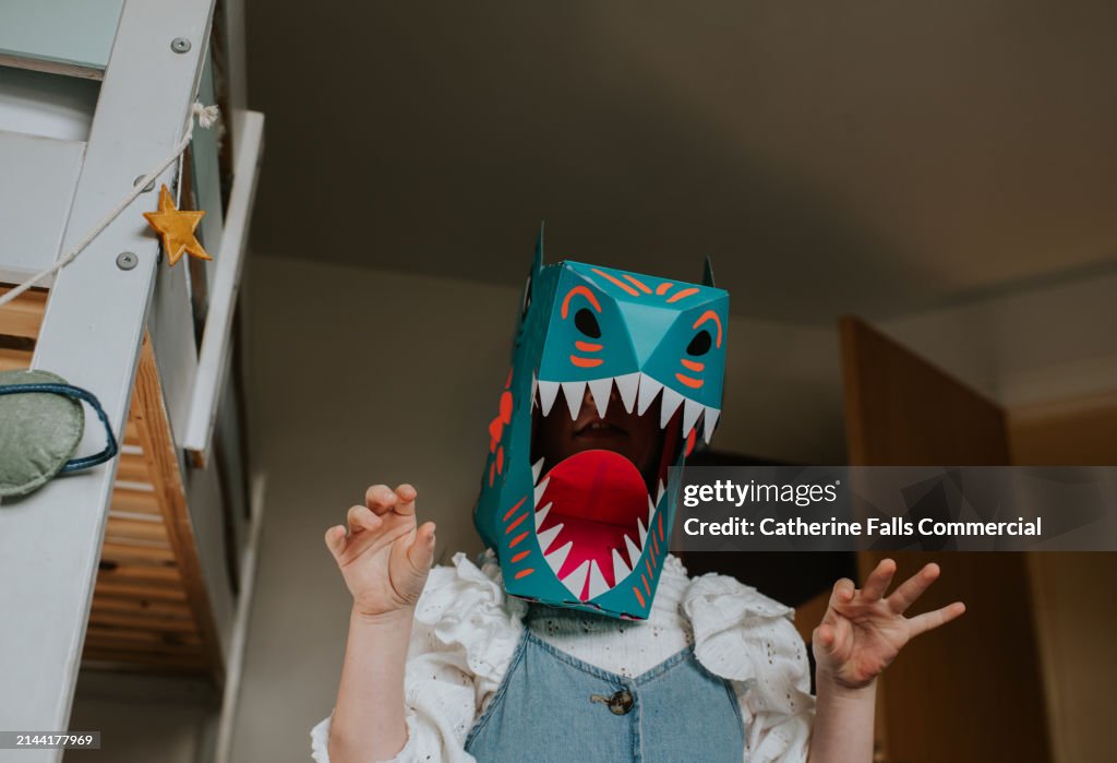 A child wears a homemade cardboard dinosaur mask, adopts a fierce pose, and roars towards the camera