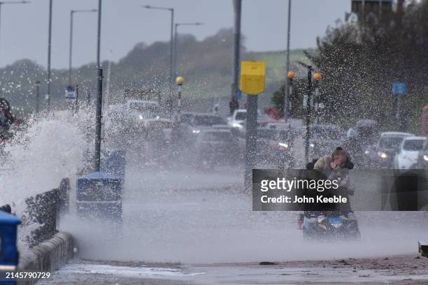 Woman gets hit with spray from a wave as she drives her mobility scooter in windy conditions along the seafront promenade in Westcliff on April 7,...
