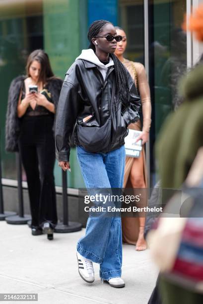 Model wears a white hoodie, a black leather jacket, blue denim jeans / pants, black and white sneakers, sunglasses, a black bag, outside La Pointe ,...