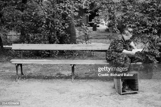 With one foot on a box, a nineties Polish woman sits on a quiet park bench reading a newspaper, on 12th July 1990, in Warsaw, Poland.