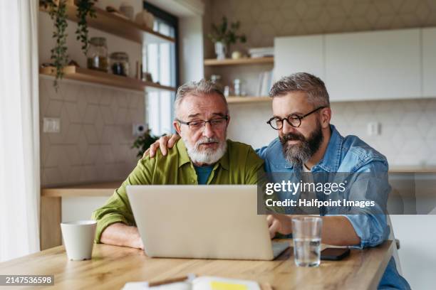 mature son helping father to manage his finance, teaching how to work with internet baking and shopping online. handsome son supporting dad, technology and digital literacy. - broma pesada fotografías e imágenes de stock