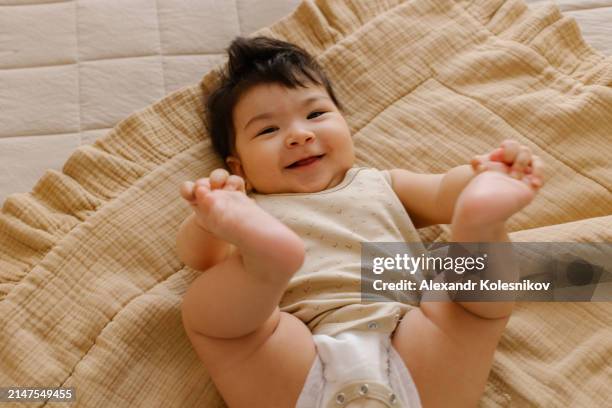 portrait of funny little baby wearing a diaper playing with his feet on a beige blanket. - supino foto e immagini stock