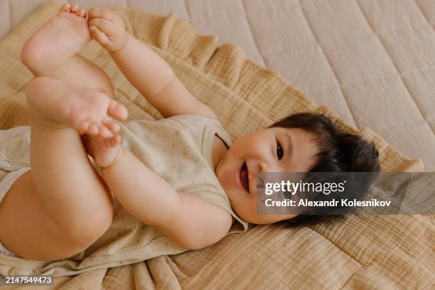 portrait of cute little smiling asian baby girl wearing bodysuit lying on beige plaid on bed at home. concept of baby's growth and development - niedlich stock-fotos und bilder