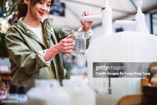 sustainable choices: a woman refilling a glass jar with biodegradable cleaning product in zero-waste store - cleaning equipment stock pictures, royalty-free photos & images