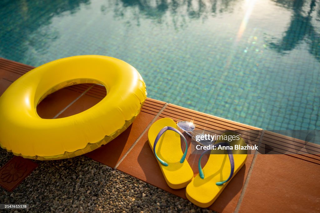 Summer flip-flops, blue sunglasses and yellow inflatable swim ring near outdoors blue pool