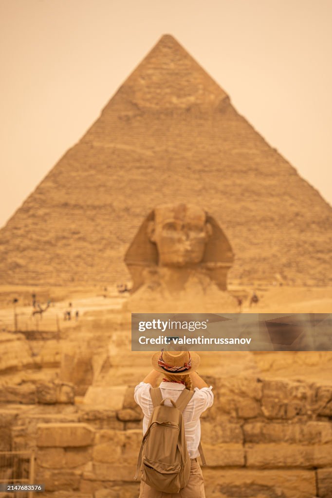 Woman standing in front of the background of Giza pyramids and Sphinx and taking pictures with smartphone. Giza, Cairo, Egypt