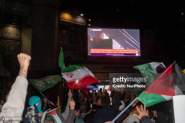 Iranians are waving flags while a display is showing a video of Iran's missile attack against Israel, during a celebration of Iran's IRGC UAV and...