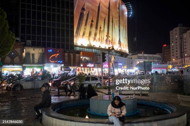 Young Iranian woman, not wearing a mandatory headscarf, is sitting at a square and using her cellphone under a massive billboard that displays an...