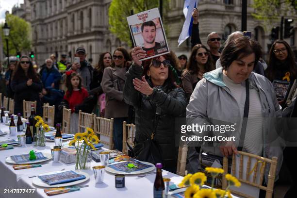 Woman holds up a poster with the photo of a young man on it as people gather around an empty seder table featuring 133 chairs representing the...