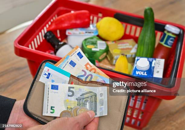 April 2024, Brandenburg, Sieversdorf: ILLUSTRATION - A woman holds money in her hand in front of a full shopping basket of groceries. The Federal...