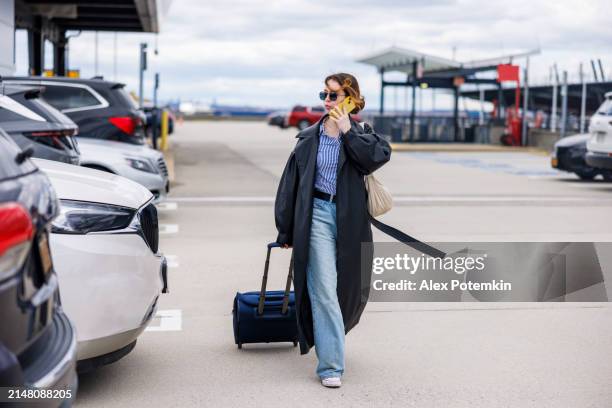 airport commute on business travel: young woman talks on the phone, pulling a suitcase on wheels through the jfk airport parking lot - john f kennedy international airport stock pictures, royalty-free photos & images