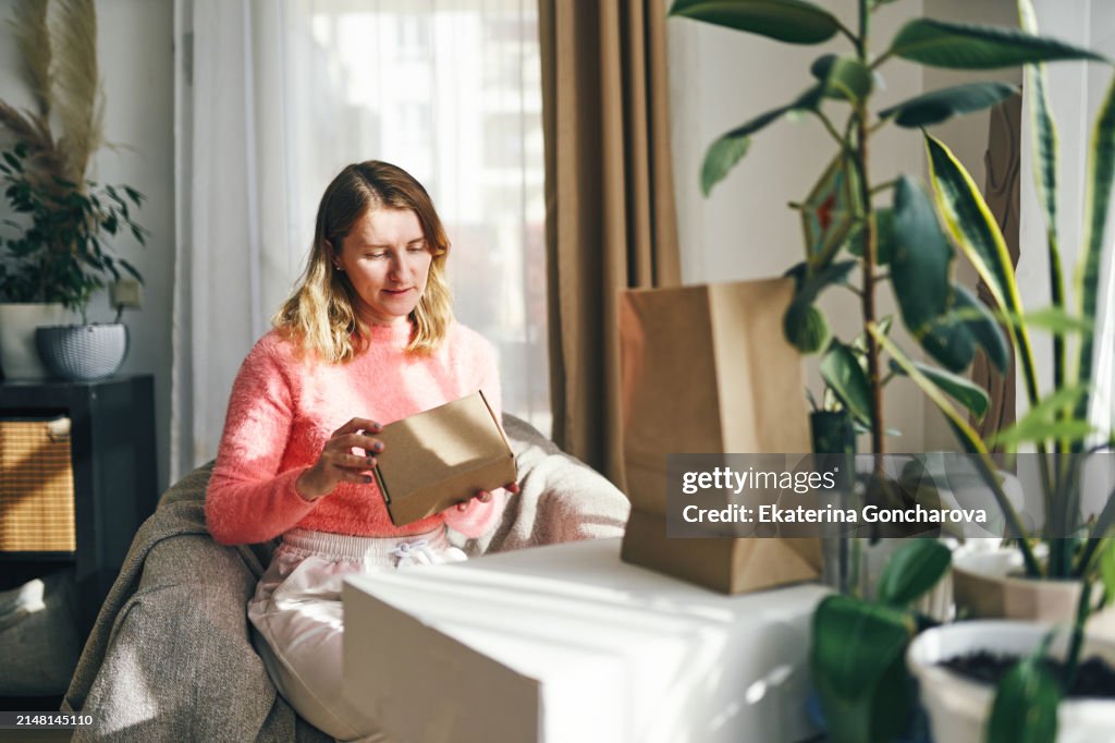 Unboxing joy. Smiling woman wrapped in a blanket on the sofa, opening a box with delight as she holds her smartphone.