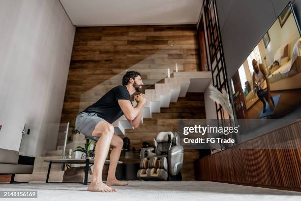 hombre adulto medio haciendo sentadillas viendo una clase de ejercicios en la televisión en casa - entrenamiento en casa fotografías e imágenes de stock