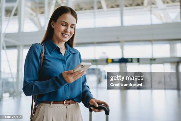 mujer en el aeropuerto sosteniendo un teléfono inteligente - mujeres de mediana edad fotografías e imágenes de stock