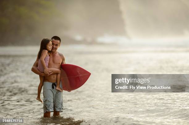 loving father carries daughter and surfboard in the ocean - pacific islanders stock pictures, royalty-free photos & images