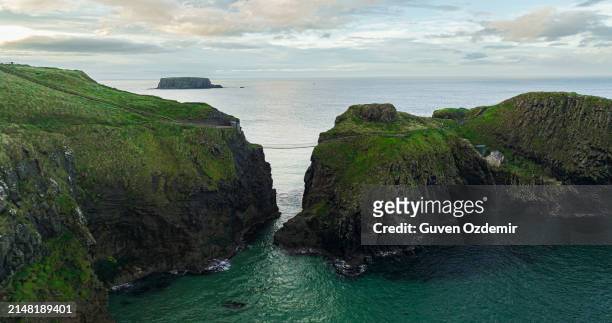 aerial view of two islands with carrick-a-rede rope bridge in northern ireland, carrick-a-rede rope bridge connecting two islands in ireland, rope bridge connecting two cliffs in northern ireland, carrick-a-rede rope bridge - hanging bridge stock pictures, royalty-free photos & images