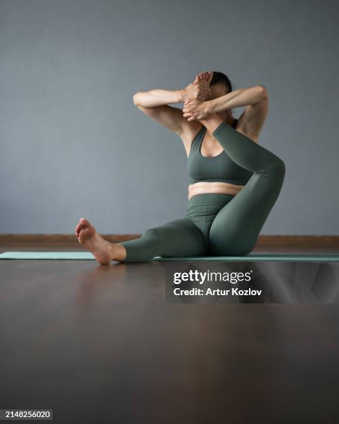 woman working out in gym on mat. eka pada shirshasana. foot against forehead. asana sitting on floor. yoga trainer. stretching. woman covers her face with her foot. front view. gray background - shirshasana stock pictures, royalty-free photos & images