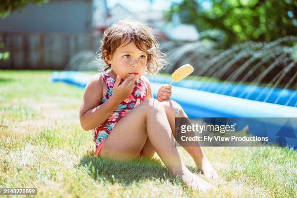 toddler girl wearing swimsuit eating popsicle in back yard - hot american girl stock pictures, royalty-free photos & images