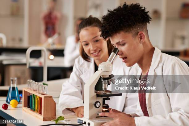 smiling teenage girl sitting by boy looking through microscope - school project stock pictures, royalty-free photos & images