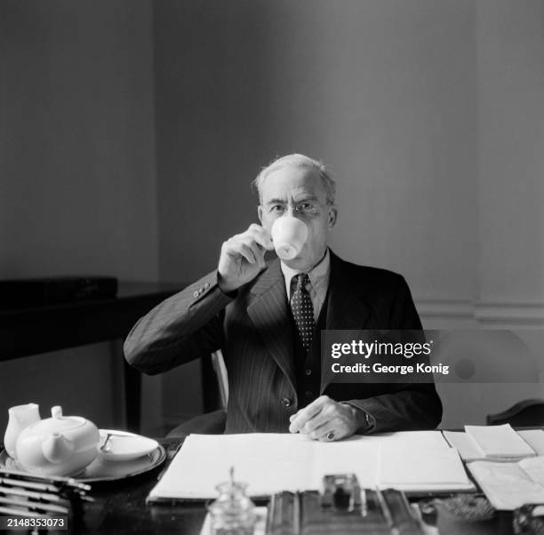 British politician Sir Stafford Cripps, the Labour Government's Chancellor of the Exchequer, drinks a cup of tea, a teapot and milk jug on a tray...