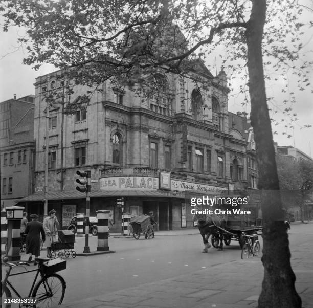 Pedestrians, with one man pulling a handcart, and a horse-drawn cart pass the Chelsea Palace Theatre on King's Road in Chelsea, West London, England,...