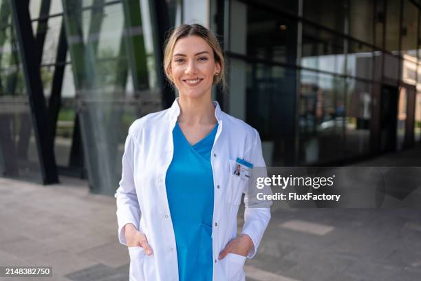 ritratto di una giovane dottoressa caucasica in camice da laboratorio e camici medici blu di fronte all'ospedale - mani in tasca foto e immagini stock