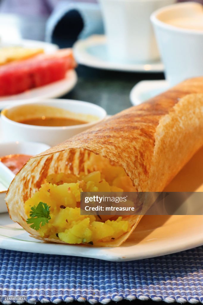 Close-up image of plate containing Indian dosa savoury pancake, crepe stuffed with mashed masala potato and chopped onions served with coconut chutney and sambar, fruit salad, tea cup and saucer, hotel restaurant table setting, focus on foreground
