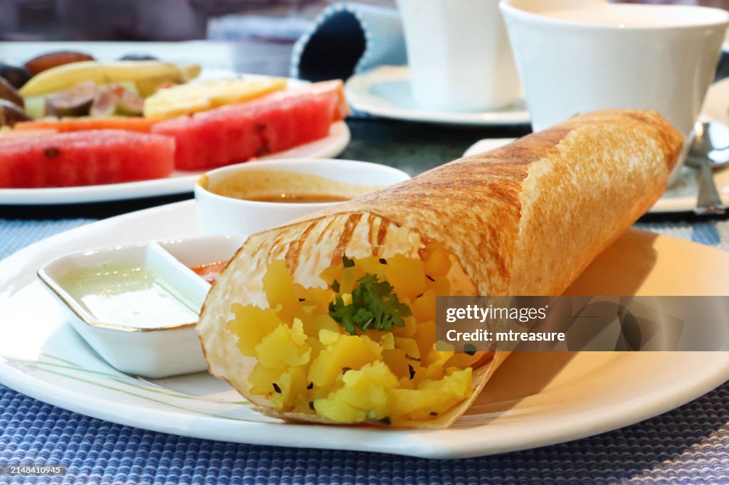 Close-up image of plate containing Indian dosa savoury pancake, crepe stuffed with mashed masala potato and chopped onions served with coconut chutney and sambar, fruit salad, tea cup and saucer, hotel restaurant table setting, focus on foreground