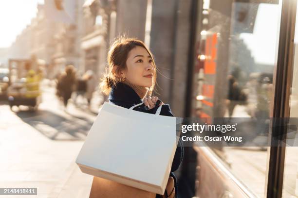 young asian woman carrying shopping bags, looking at store window - ein tag im leben stock-fotos und bilder