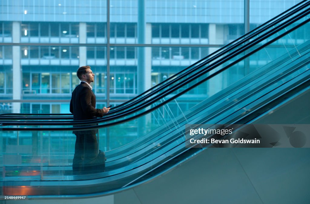 Business, commute and man on escalator in office with phone, opportunity and ambition in professional job. Travel, thinking and businessman in lobby with window, smartphone and luggage for work trip