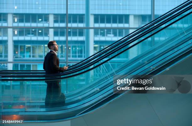 business, commute and man on escalator in office with phone, opportunity and ambition in professional job. travel, thinking and businessman in lobby with window, smartphone and luggage for work trip - potential stock pictures, royalty-free photos & images