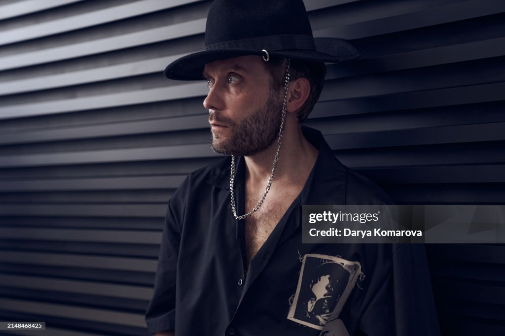 Close up portrait of a handsome man in a black shirt and pierced hat is sitting on the black background. Men with beard, perfume concept with copy space.